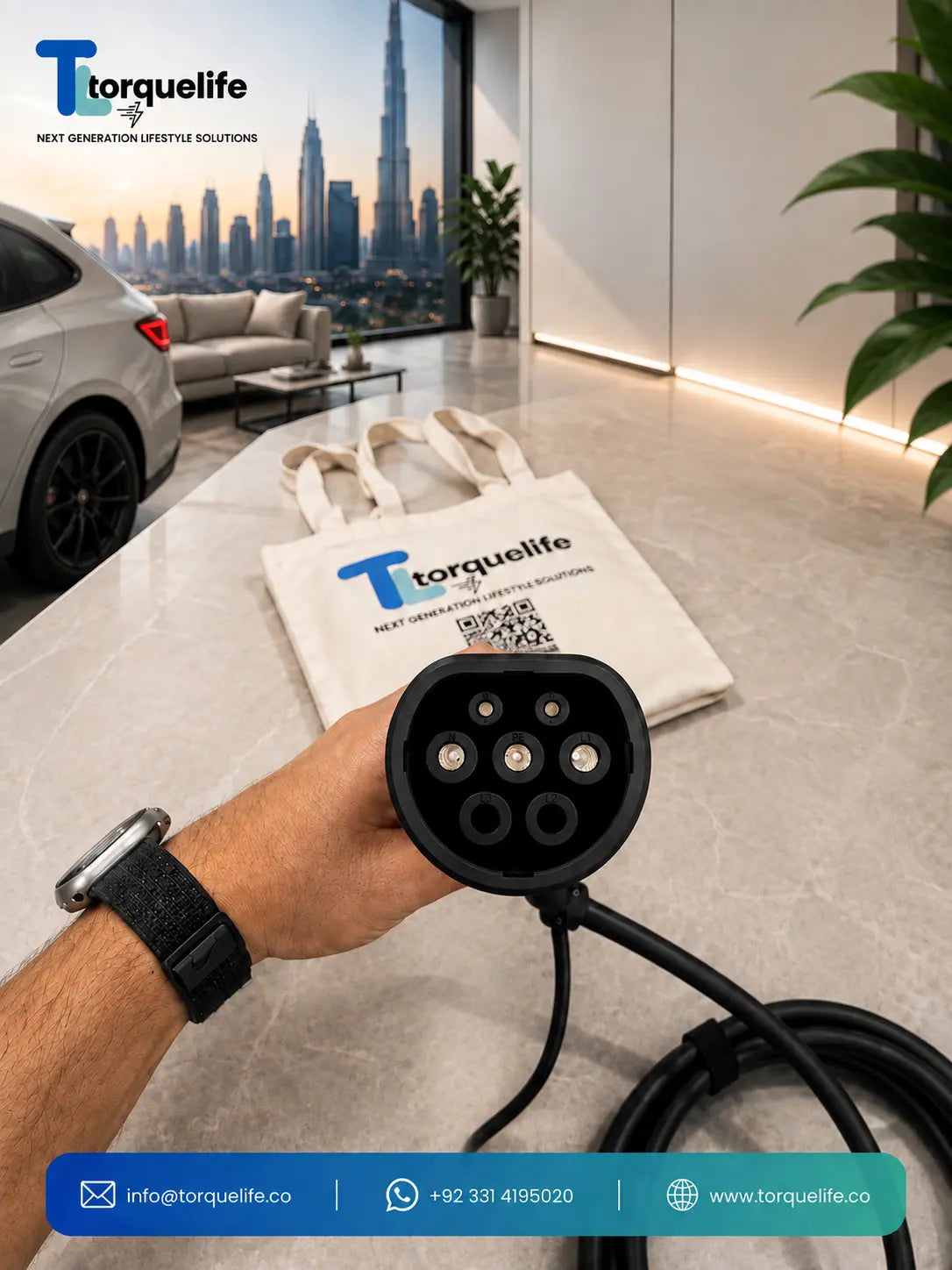 A hand holds a black electric vehicle charging connector type 2 with its attached cable in a modern office or showroom setting. A branded white tote bag with the Torquelife logo sits on the counter behind it, with a car and city skyline visible in the background.